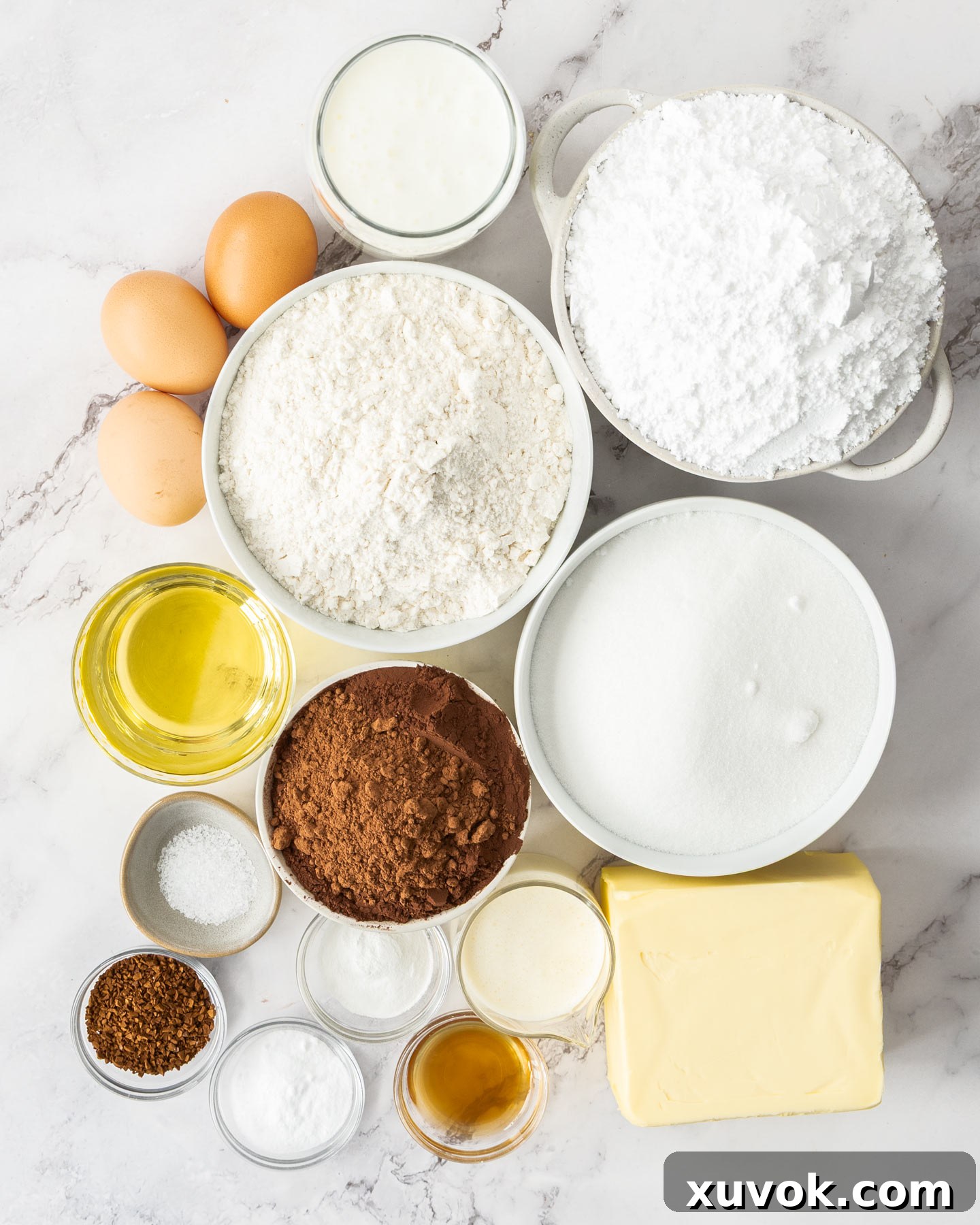 A collection of ingredients laid out on a white surface, including cocoa powder, flour, eggs, butter, and vanilla extract, ready for baking the chocolate cake with vanilla buttercream.