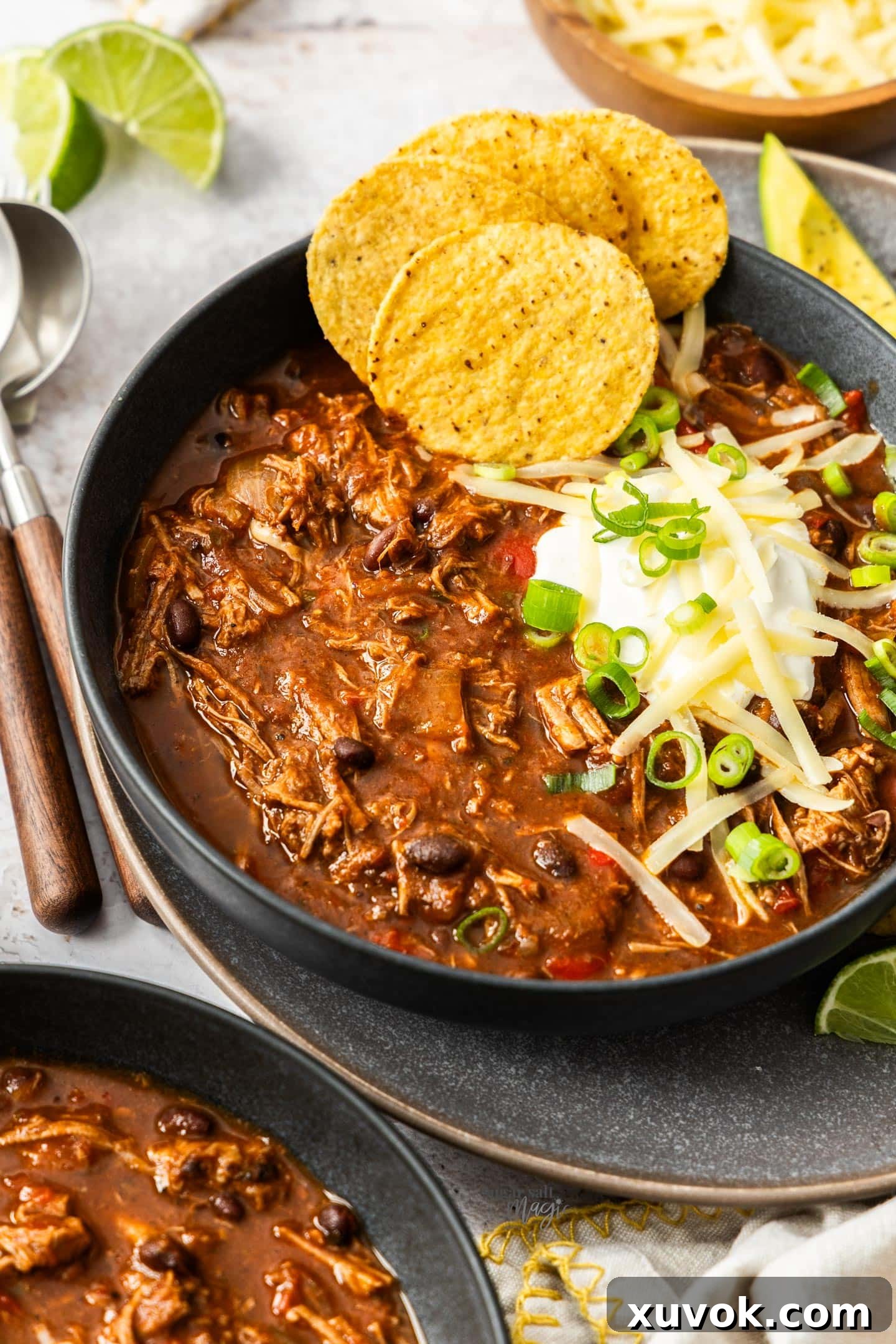 A dark bowl filled with rich chilli con carne, garnished with a sprinkle of cheese and a side of crispy corn chips.