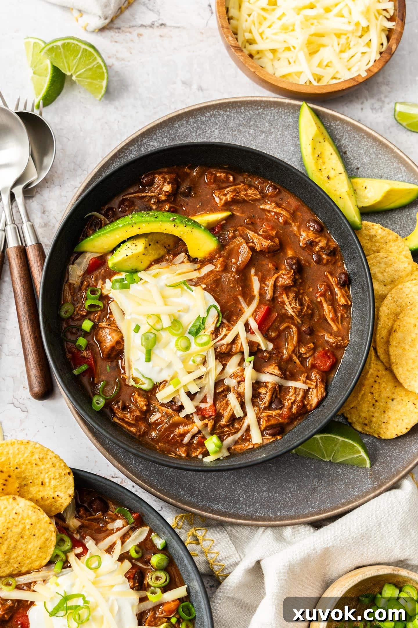 Top-down view of a dark bowl brimming with slow cooker chilli con carne, garnished generously with fresh avocado slices, cool sour cream, and shredded cheese.