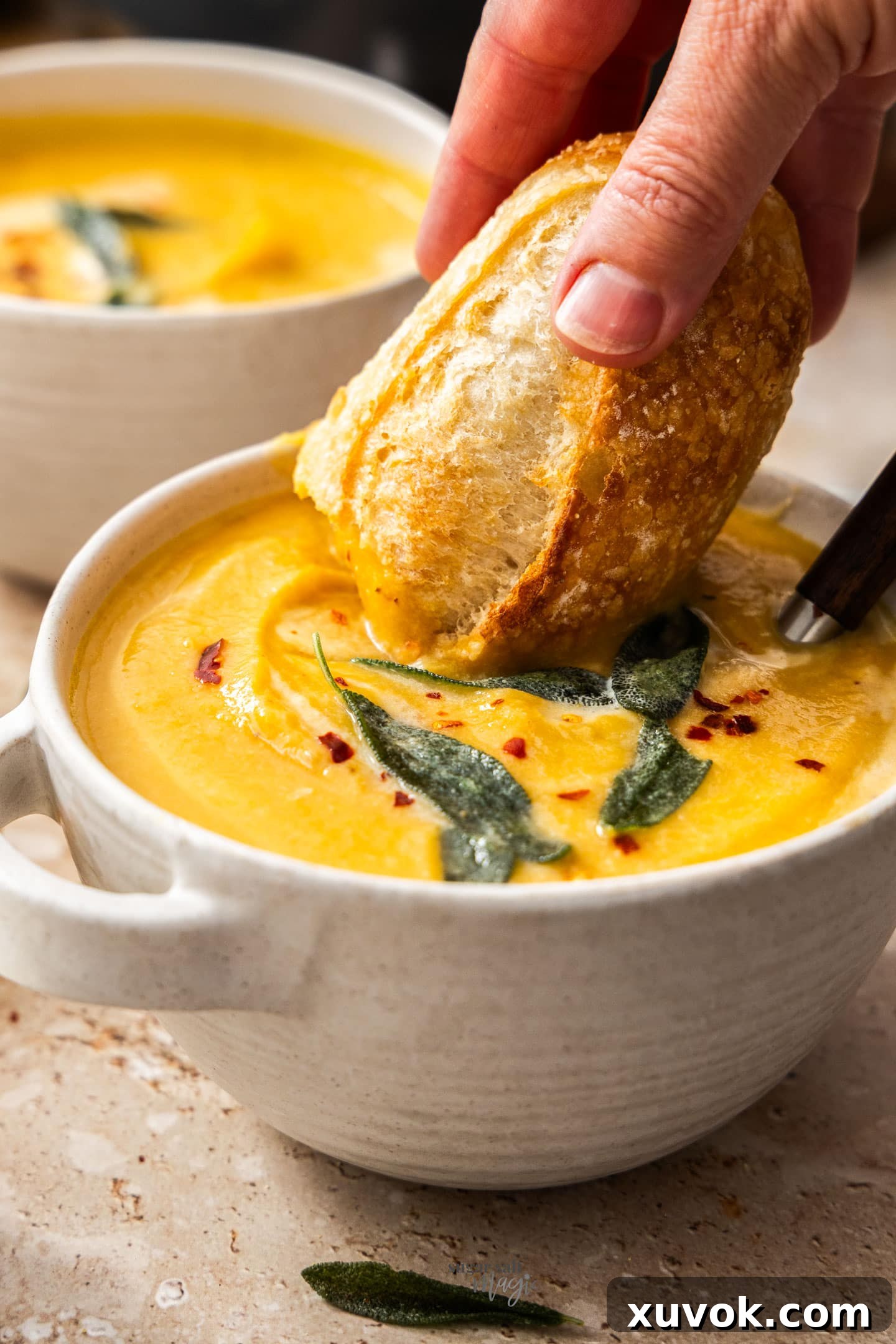 A chunk of rustic bread being dipped into a rich, creamy bowl of cauliflower and pumpkin soup.