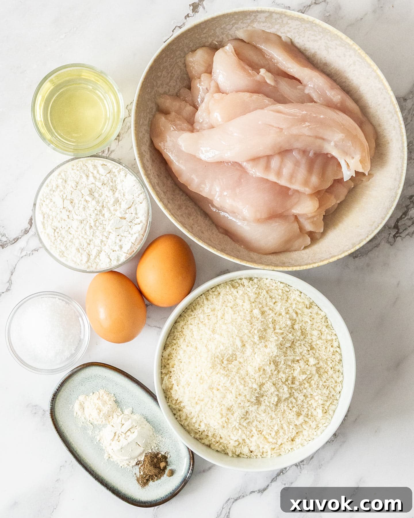 Top down view of ingredients for crumbed chicken tenders on a marble surface.