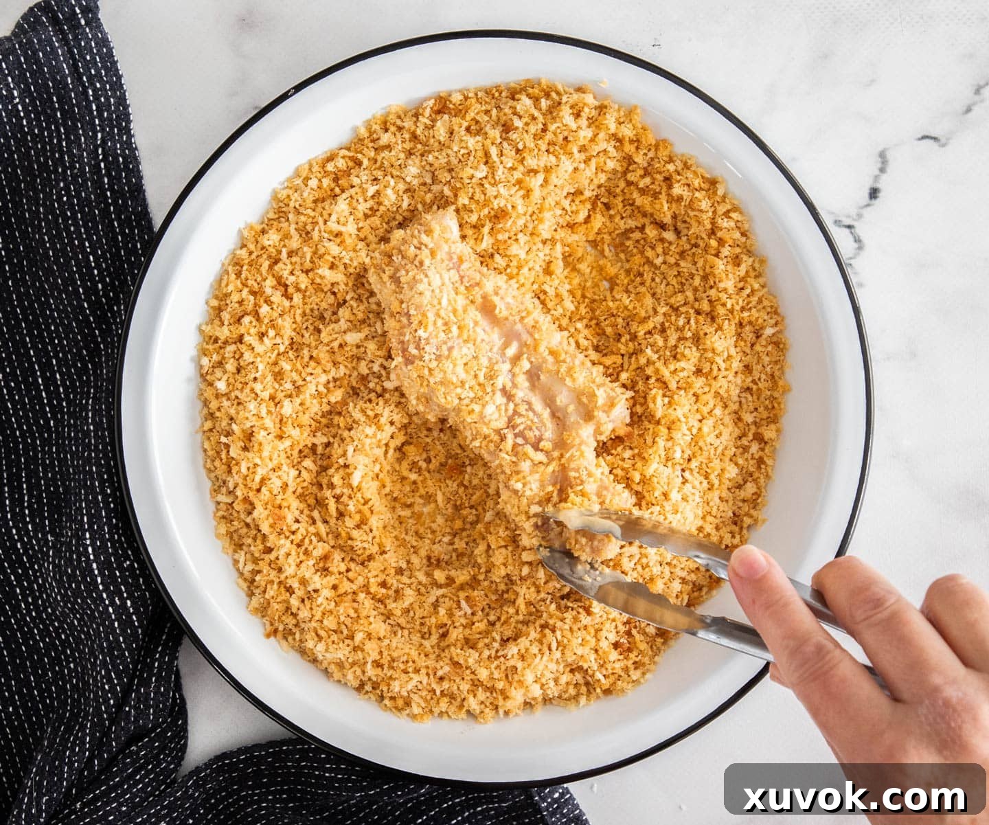 A hand using tongs to coat the chicken in crumbs.