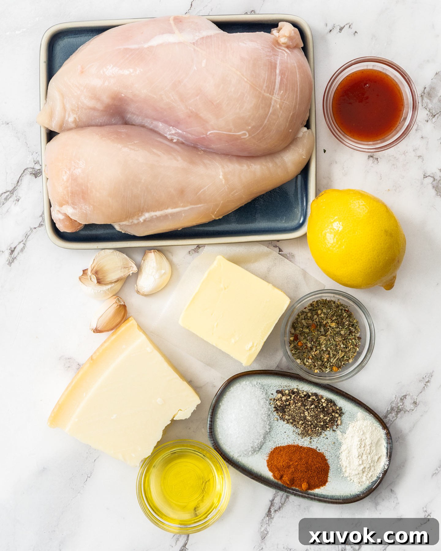 Top down view of fresh ingredients for garlic parmesan chicken skewers on a marble surface, including chicken, butter, garlic, lemon, and spices.