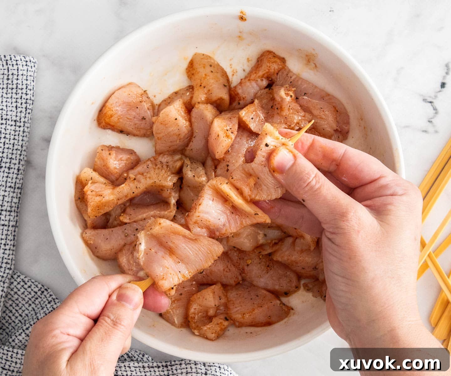 Two hands skillfully pushing chunks of seasoned chicken onto wooden skewers.
