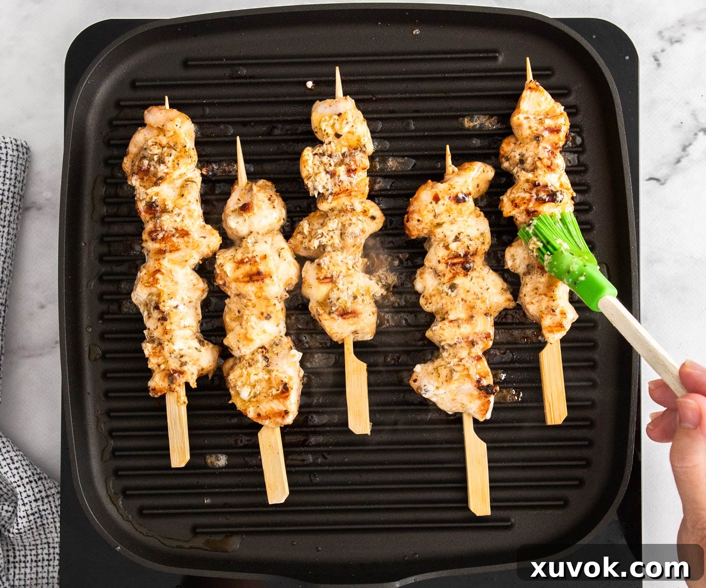 Chicken skewers actively cooking and being basted with a savory butter sauce on a griddle pan.