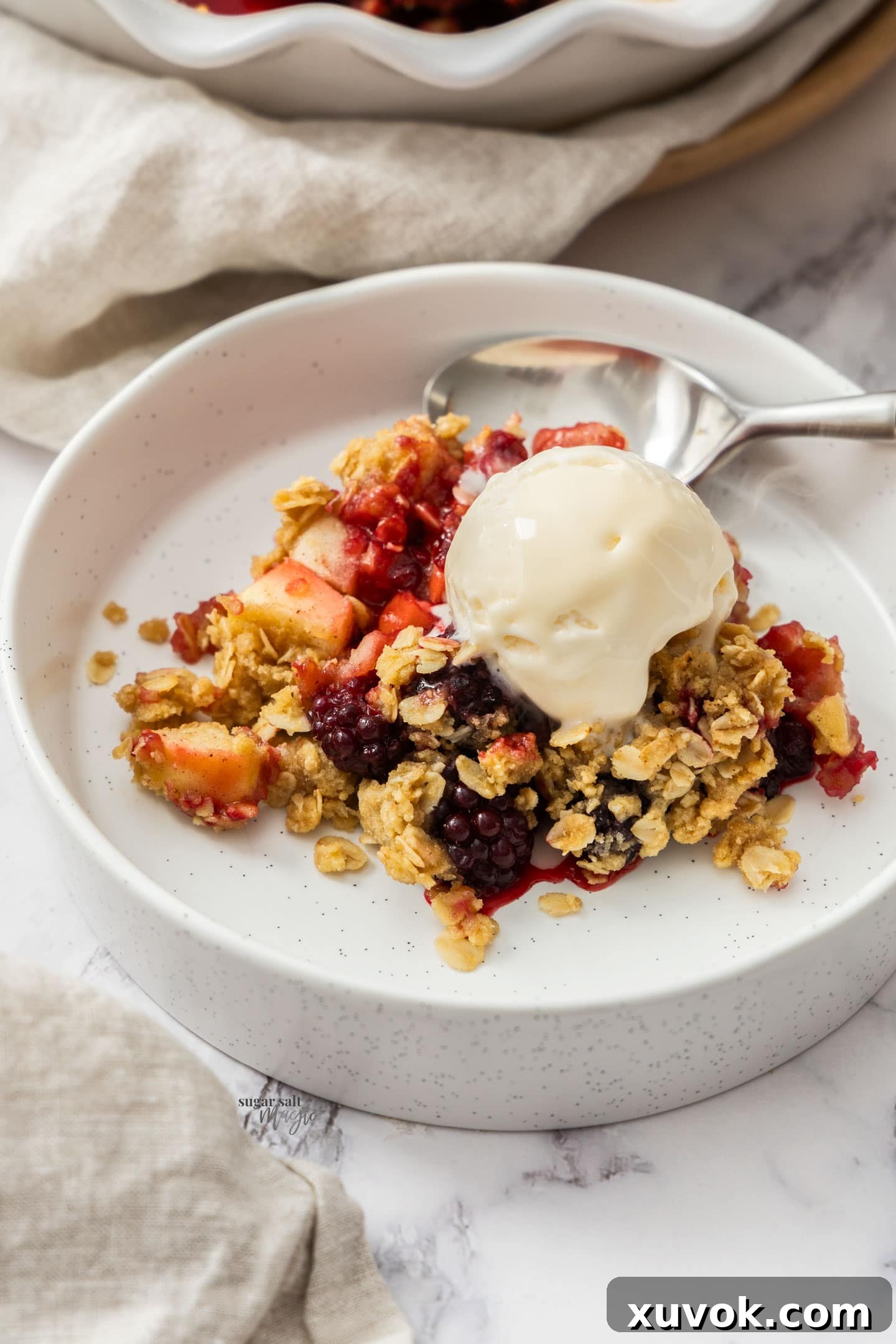 A close-up view of a bowl of apple and blackberry crumble, showing the perfectly cooked fruit peeking through the golden, crunchy topping, garnished with a melting scoop of ice cream.