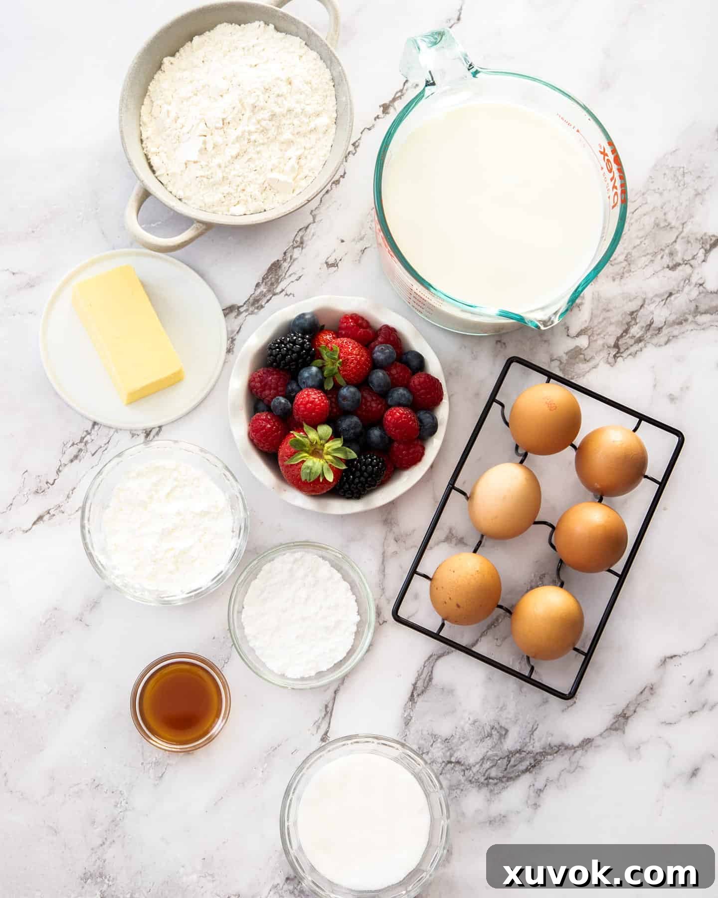 A collection of fresh ingredients for fruit custard tart, including flour, butter, sugar, eggs, milk, cornflour, vanilla extract, and a variety of berries, laid out on a marble worktop.