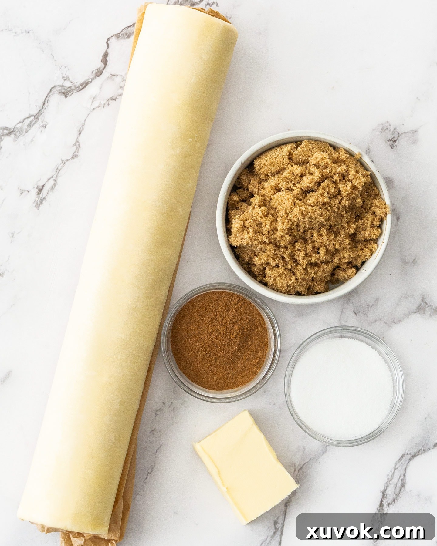Ingredients for puff pastry cinnamon twists laid out on a marble countertop, including puff pastry, butter, sugars, cinnamon, and an egg.