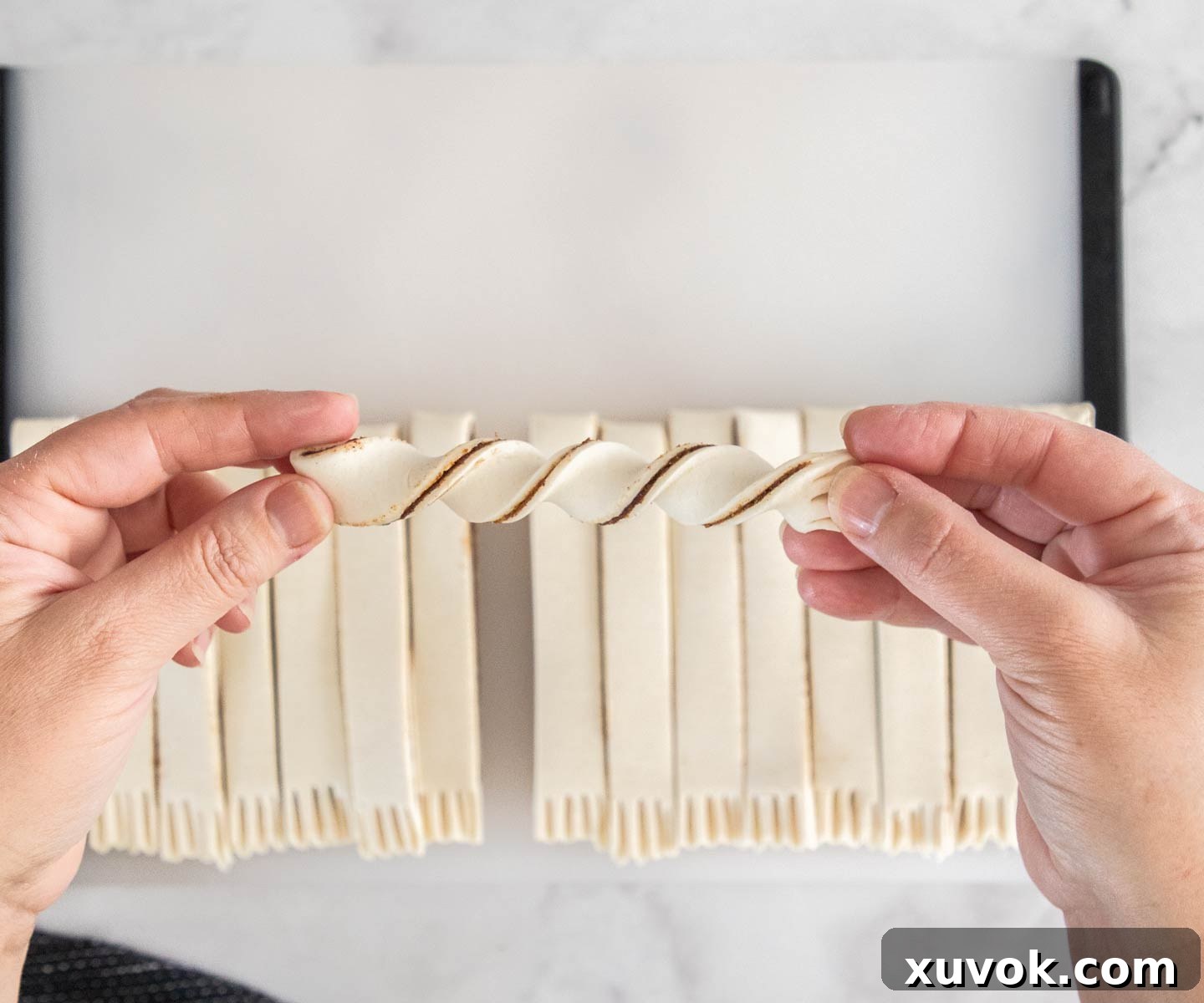 A pair of hands demonstrating how to twist a raw cinnamon puff pastry strip into its characteristic spiral shape.
