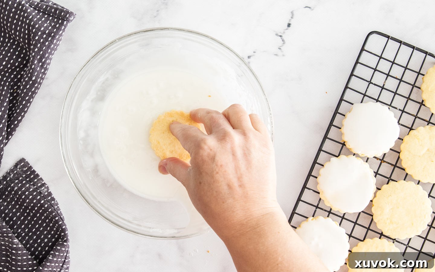 A hand dipping a baked cookie into the almond icing, illustrating the glazing process.
