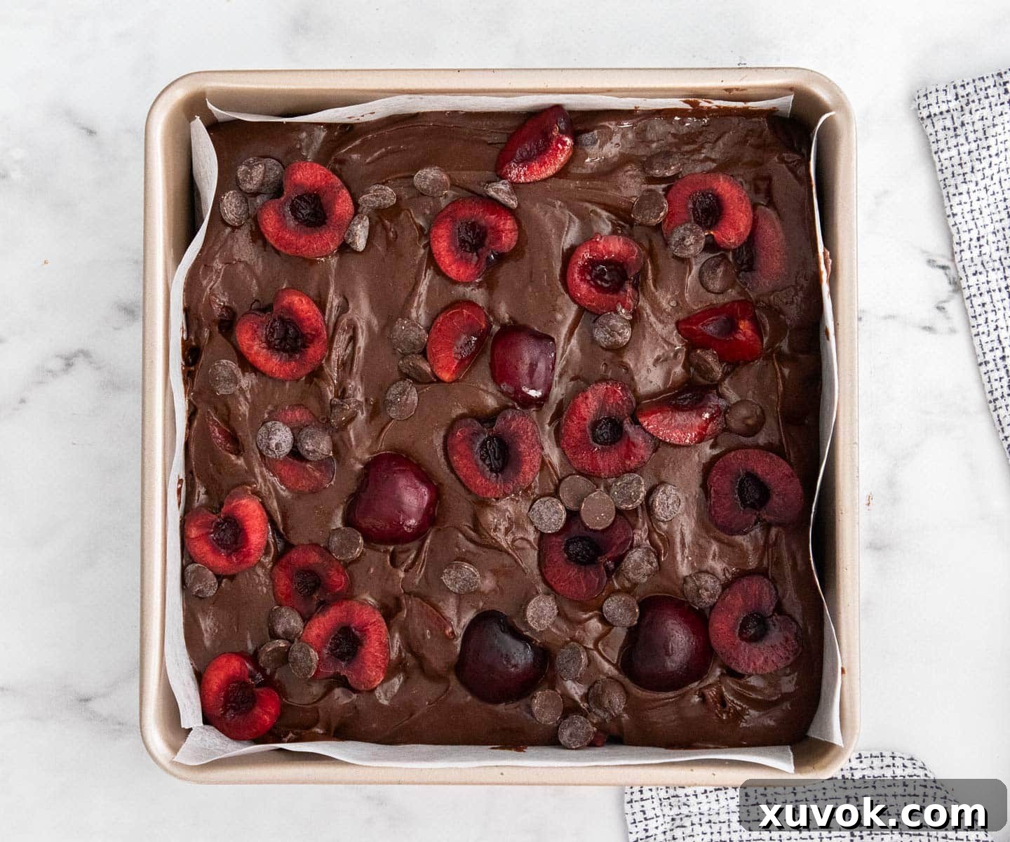 The cherry brownie batter in a baking pan, generously topped with fresh cherries and chocolate chips, ready for baking.
