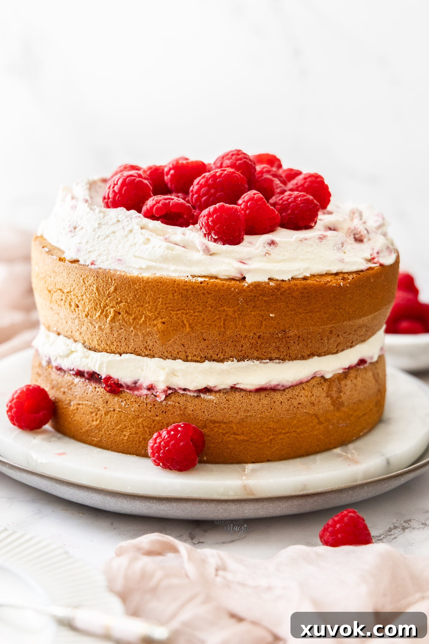 A two-layer raspberry sponge cake on a marble platter, piled high with fresh raspberries and Chantilly cream.