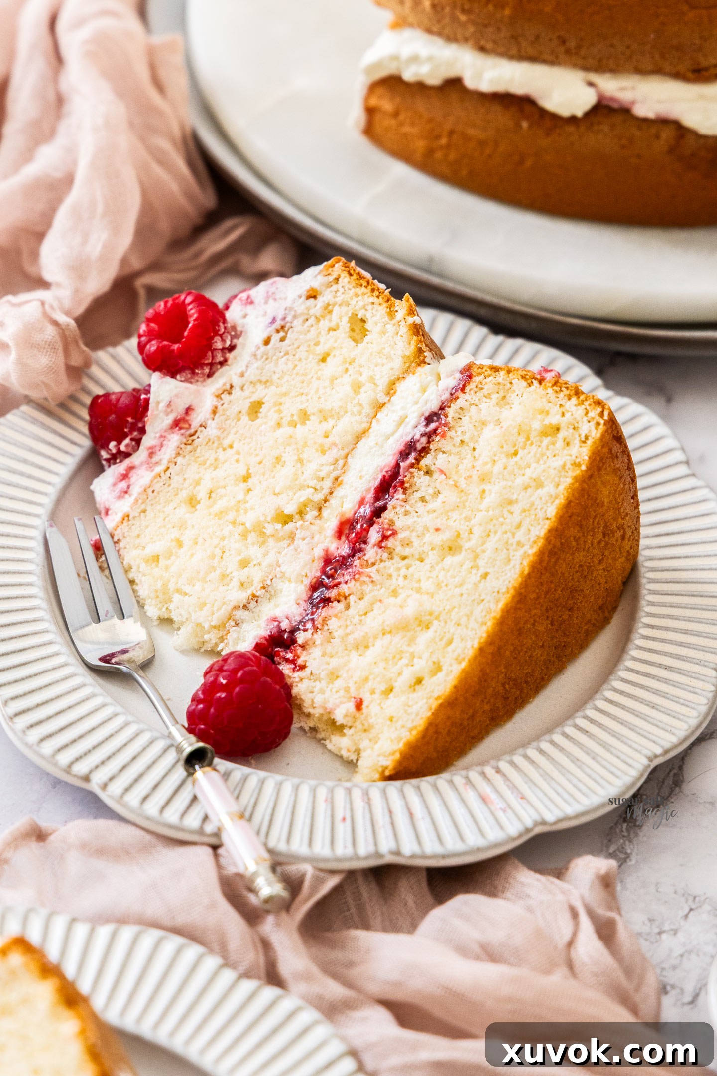A perfectly sliced piece of raspberry sponge cake with layers of cream and jam, served on a white plate with a dessert fork.
