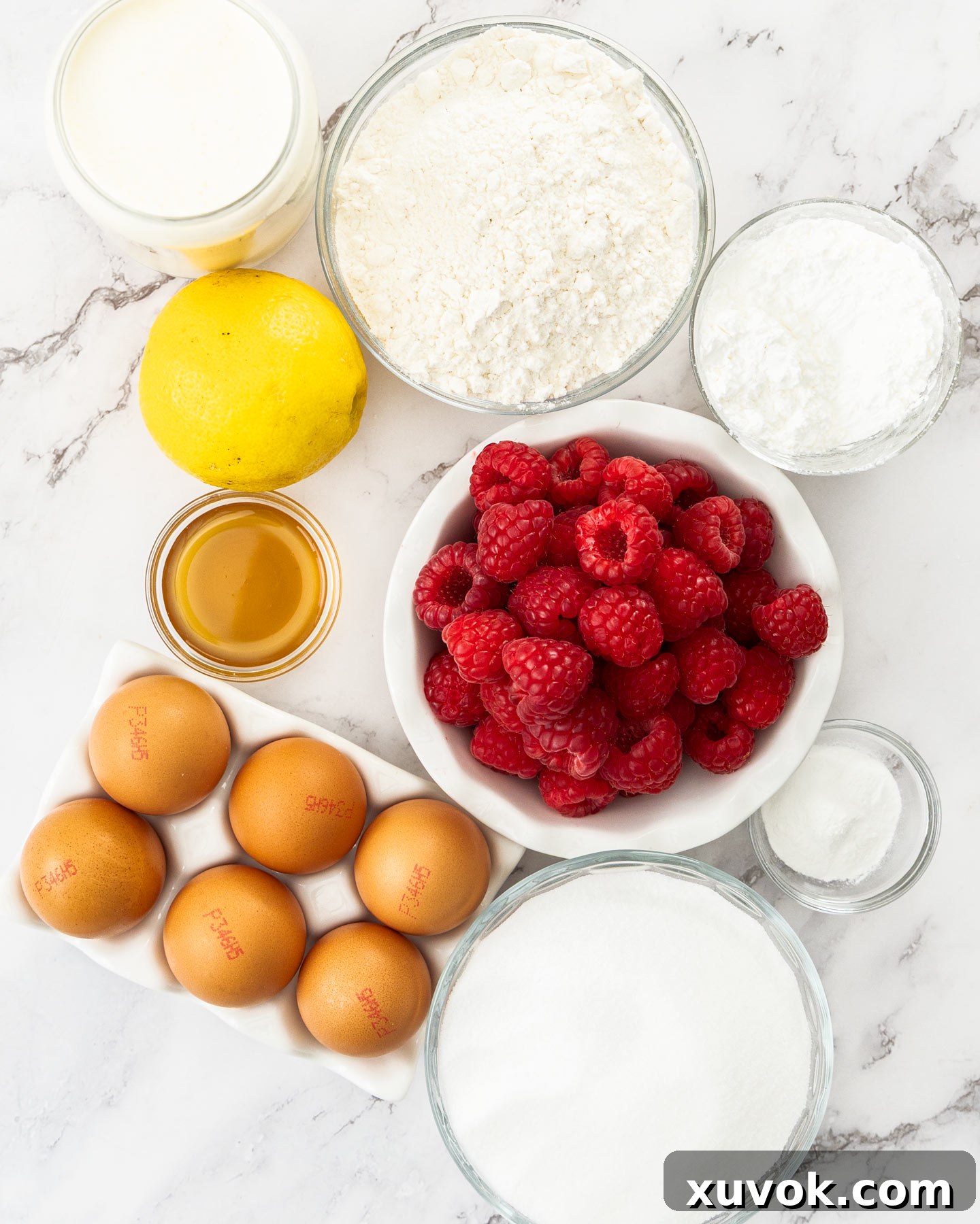 A flat lay photograph displaying all the ingredients required for making a raspberry sponge cake, including fresh raspberries, eggs, flour, sugar, and cream.