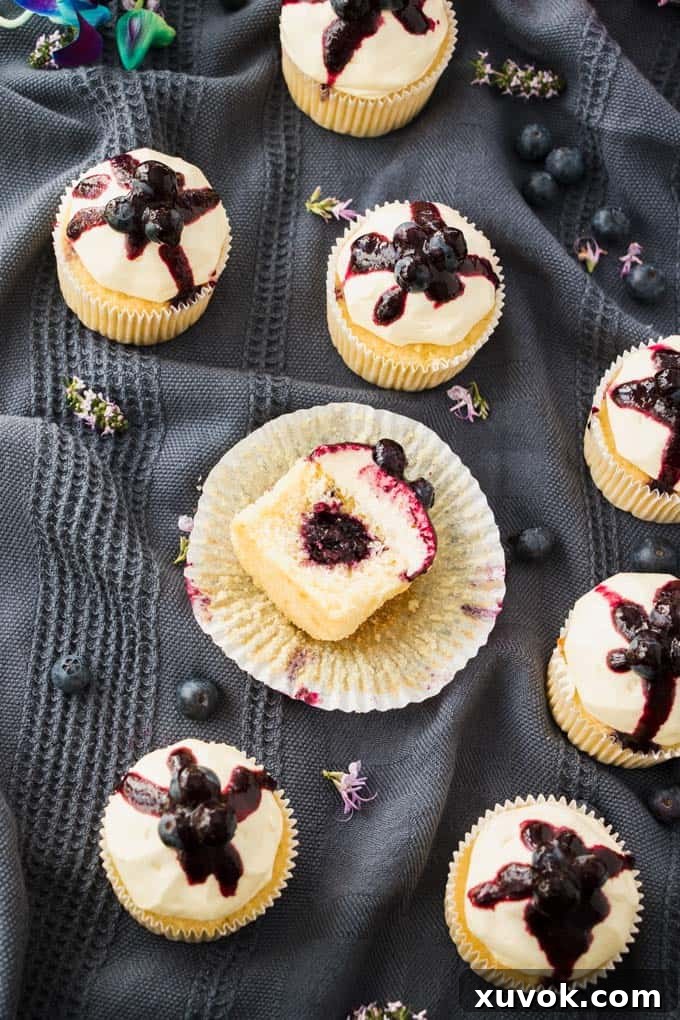 A Lemon Blueberry Cupcake artfully cut in half, revealing the delightful blueberry compote center, surrounded by fresh blueberries and more cupcakes.