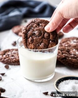 A chocolate cookie being dunked into a glass of milk with more cookies scattered around it