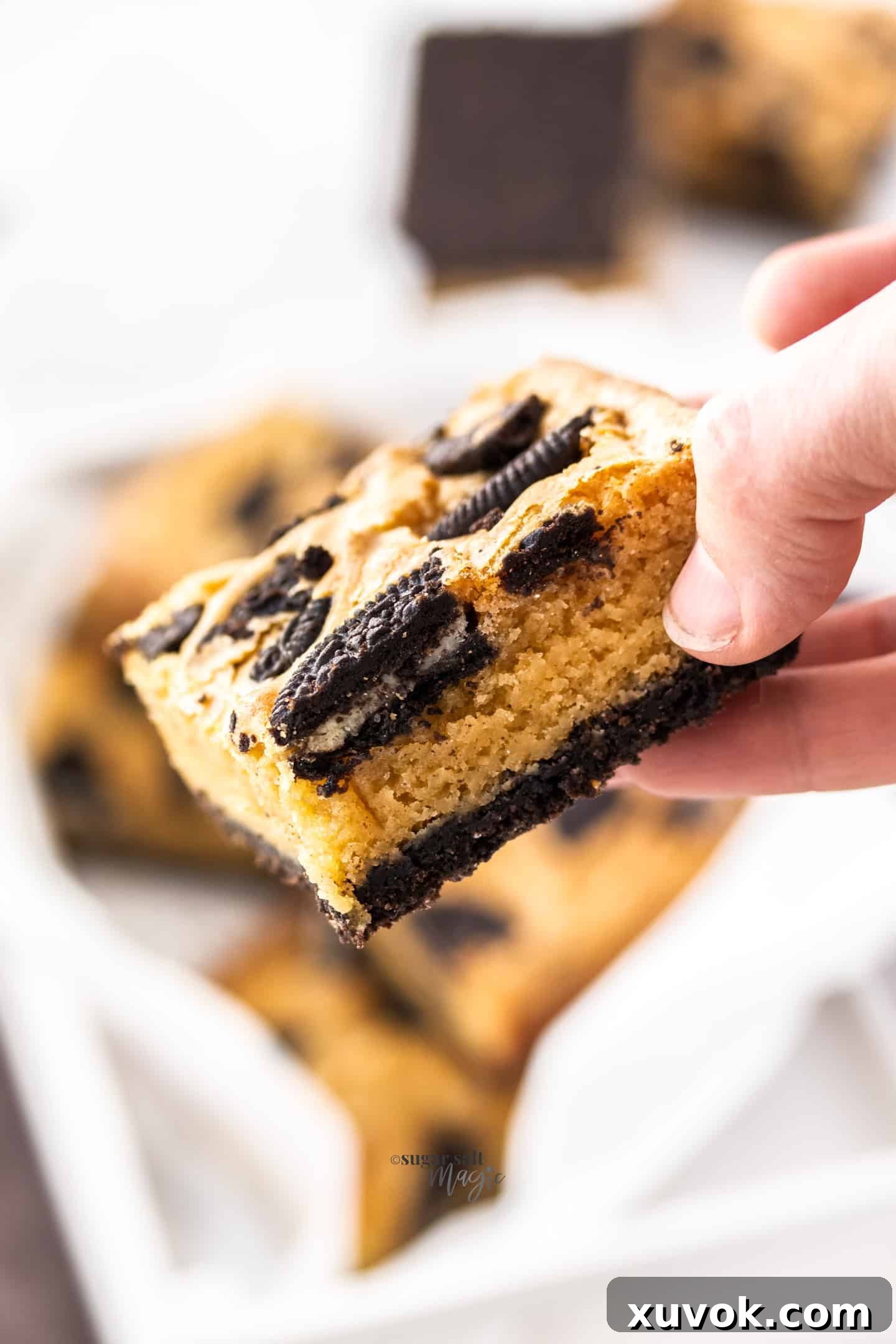 A close-up of a hand holding a perfectly baked Oreo blondie, broken in half to reveal its dense, fudgy interior and the distinct layers of the crunchy Oreo base and cookie topping.
