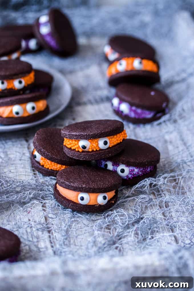 A pile of 4 Chocolate Monster Halloween Cookies with more in the back and on a plate. Each cookie features colorful buttercream and playful candy eyes.