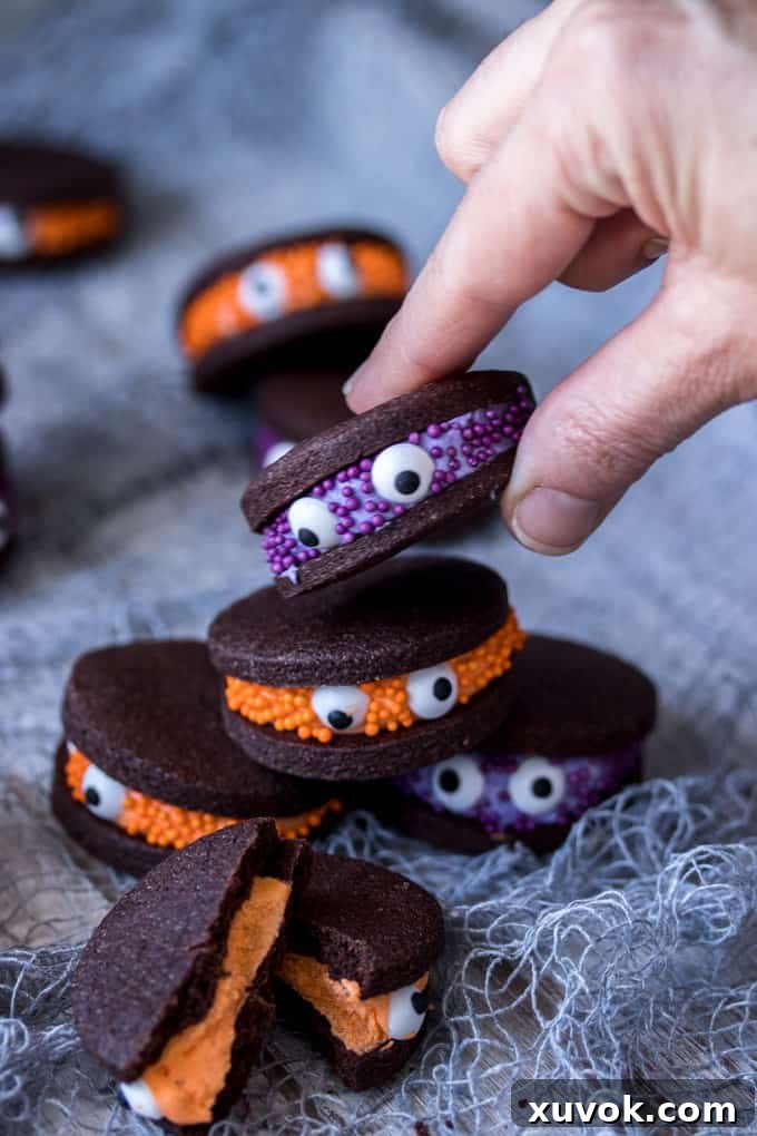 A hand picking up a chocolate sandwich cookies with purple filling, decorated with purple sprinkles and eye balls. The cookie looks playful and inviting.
