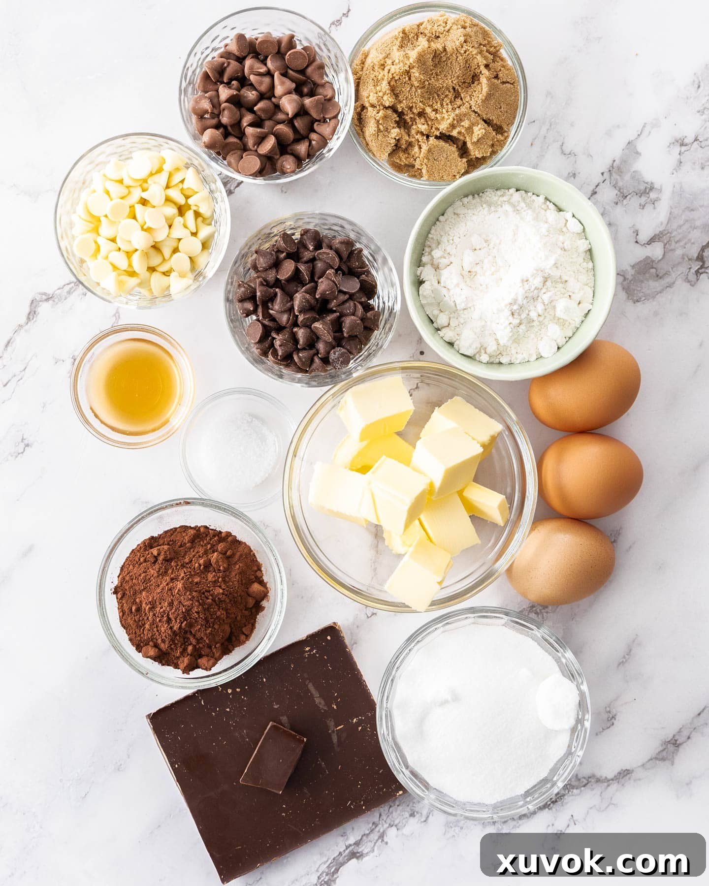 Ingredients for triple chocolate brownies laid out on a marble surface, including butter, chocolate, eggs, sugars, flour, cocoa, and various chocolate chips.