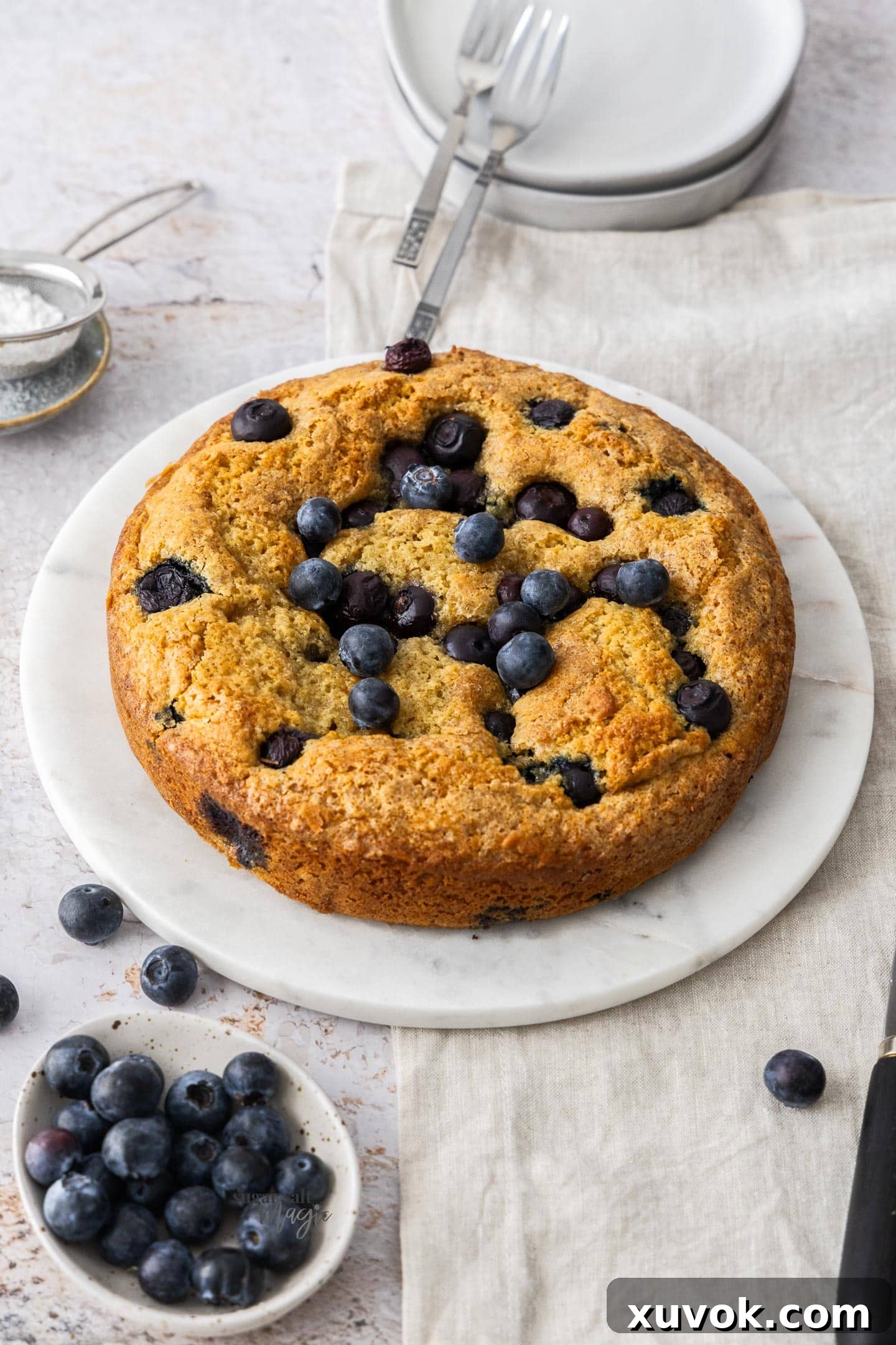 A whole blueberry cake on a platter with a small bowl of blueberries in front.