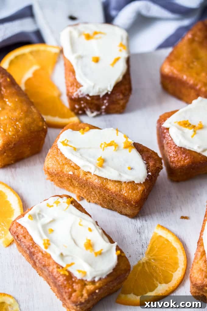 A few Mini Flourless Ginger Orange Cakes, some with white frosting, on a white board, next to orange slices. Perfect for a dessert table.