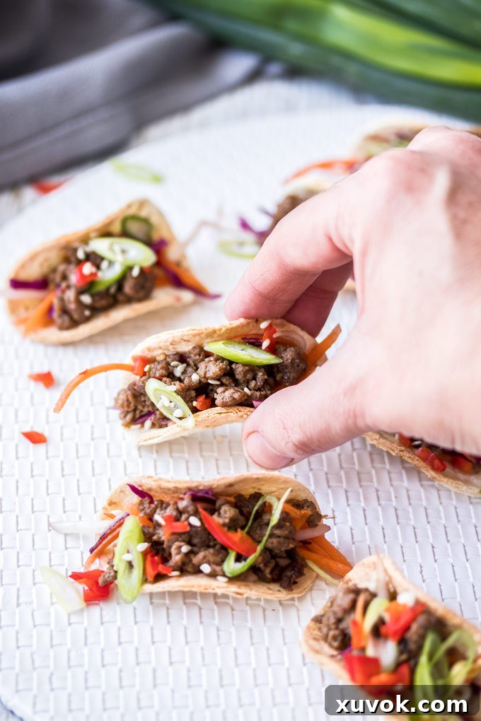 A hand reaching, picking up a mini korean beef taco off a white mat with others around it and green onions in the background. The taco is filled with ground beef bulgogi and Asian slaw.