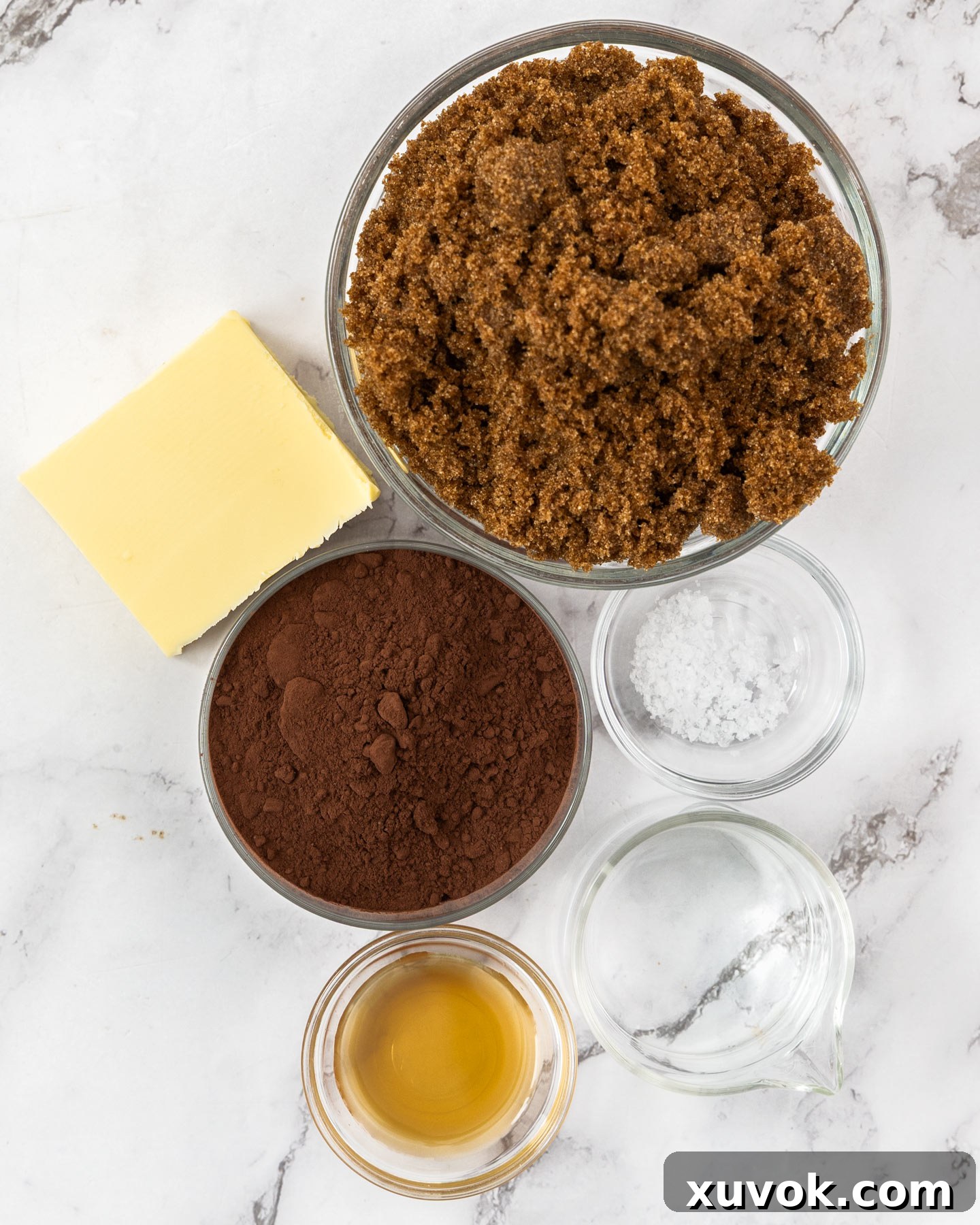 Ingredients for homemade chocolate sauce laid out on a marble background, including cocoa, sugar, butter, and vanilla.