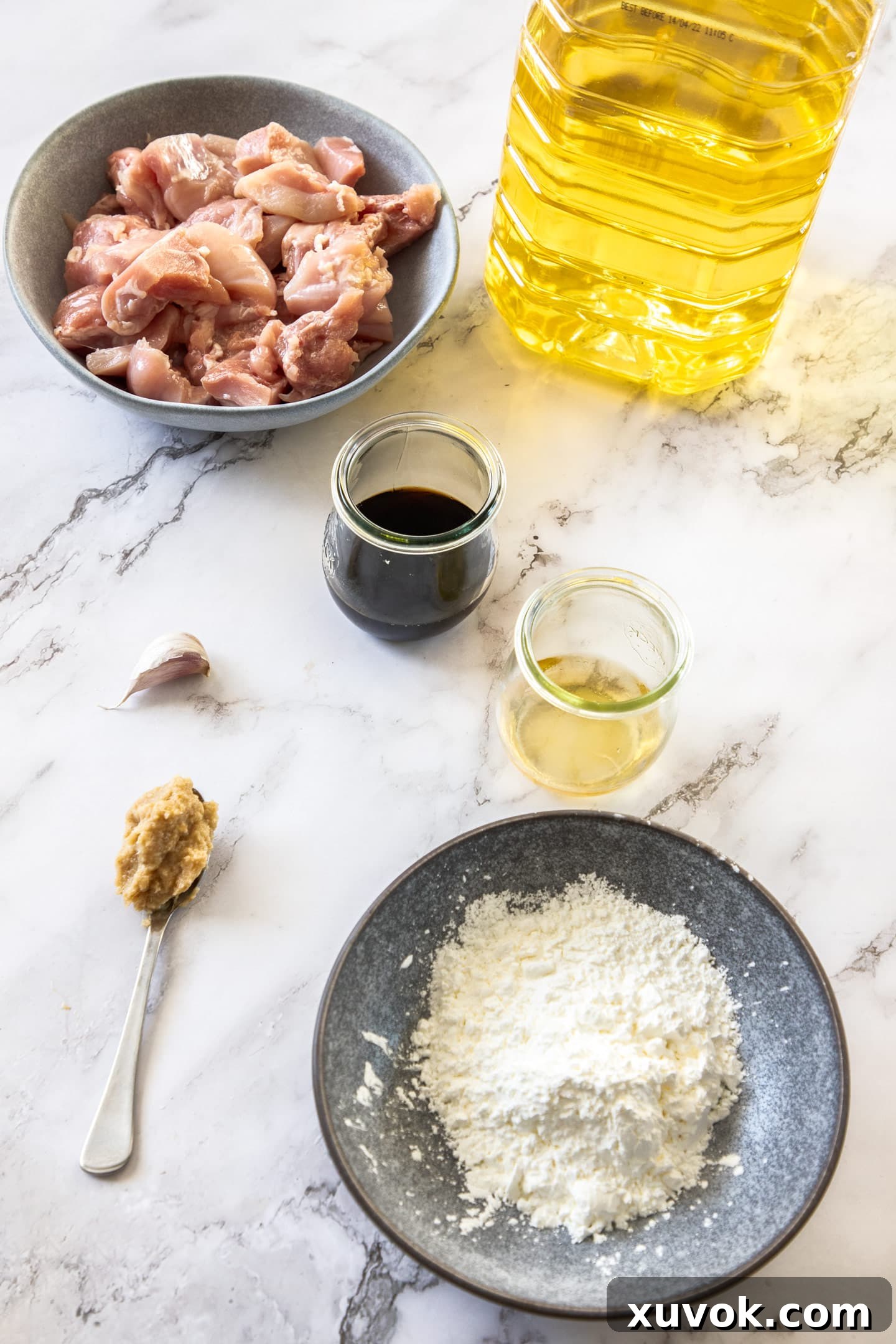 A vibrant flat lay showcasing all the essential ingredients for Karaage Chicken, including chicken thighs, soy sauce, mirin, potato starch, fresh ginger, garlic, and cooking oil, neatly arranged on a white marble surface.