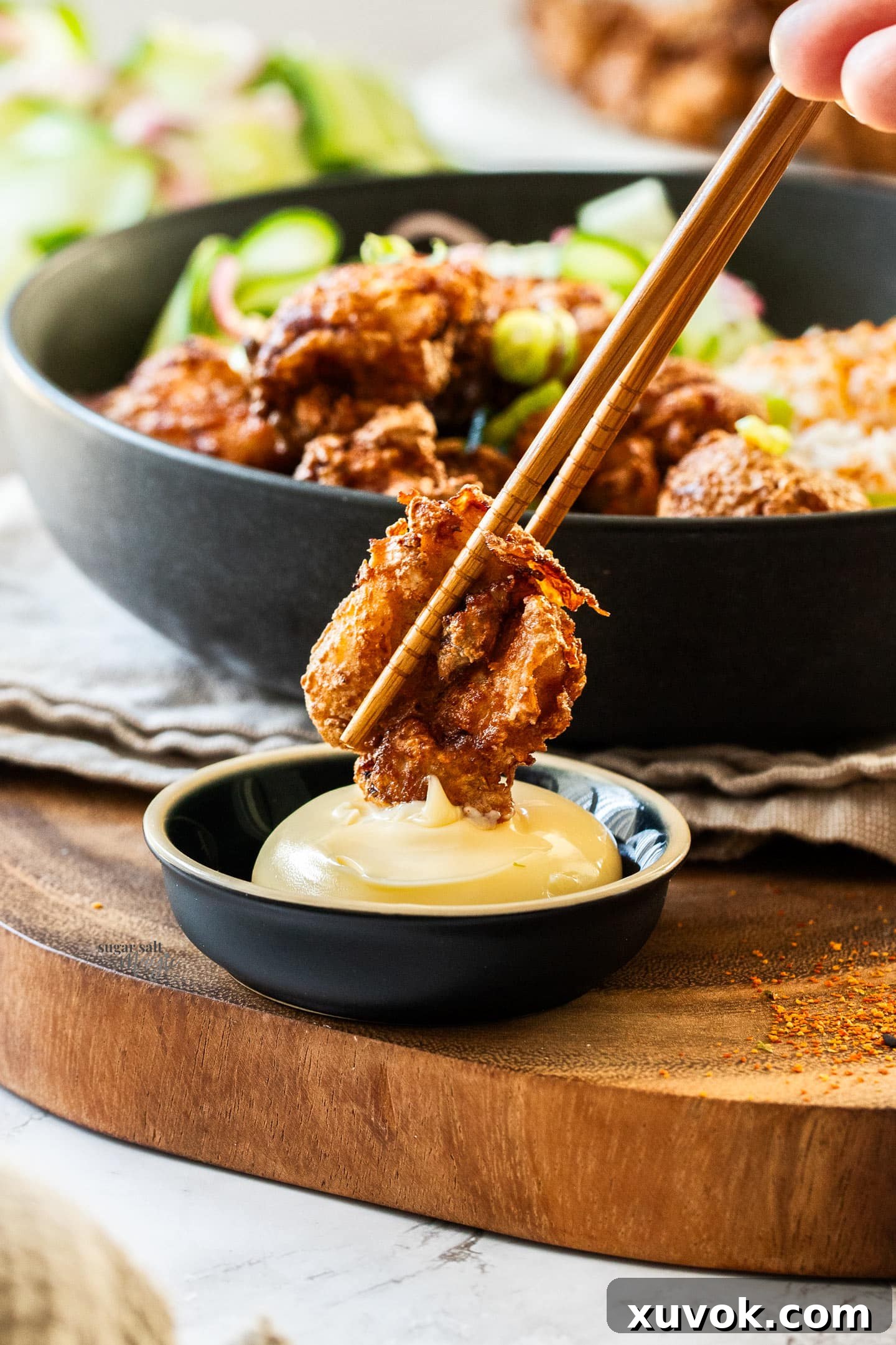A pair of chopsticks delicately holding a piece of golden-brown Karaage chicken, dipping it into a small white bowl of creamy Kewpie mayonnaise.