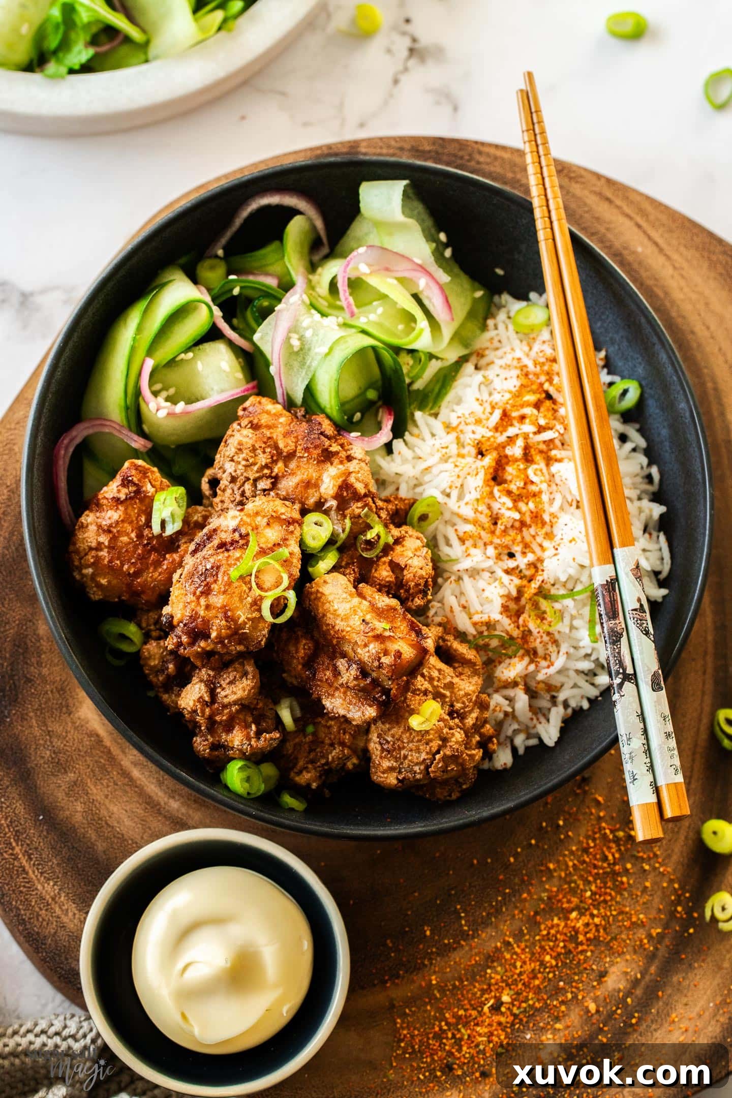 A top-down view of a beautifully arranged Japanese meal featuring golden-brown Karaage chicken in a bowl with fluffy white rice and sliced cucumber, all on a rustic wooden board.