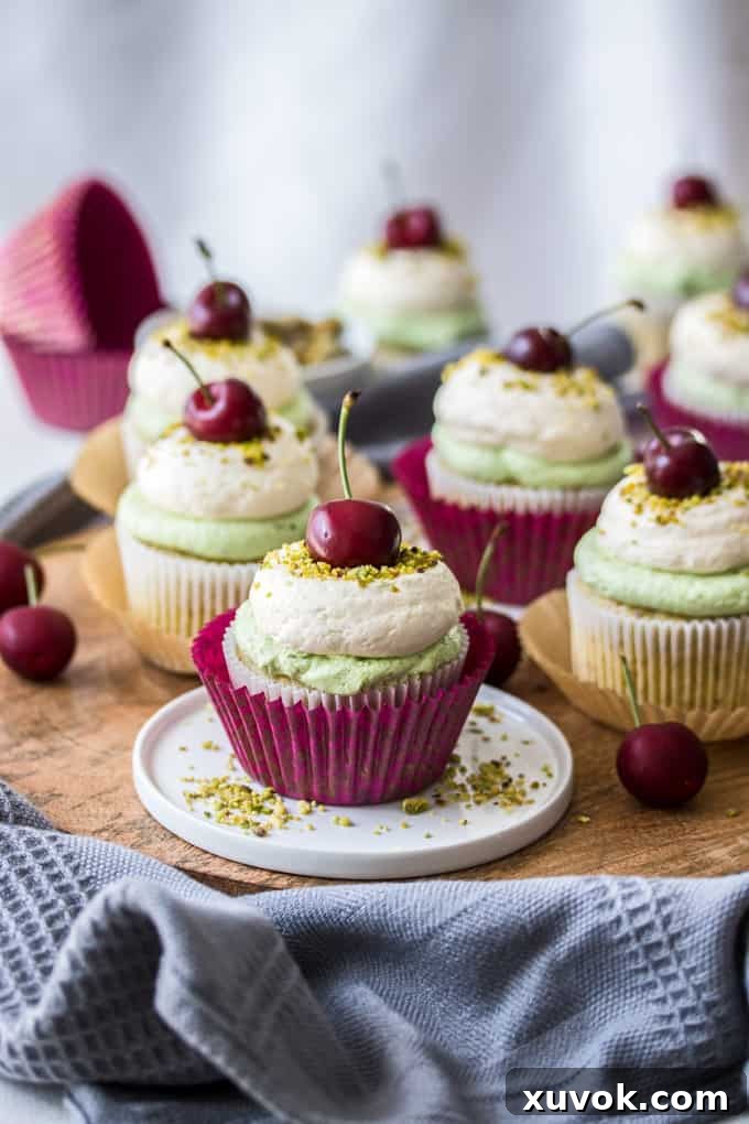 A single Cherry Pistachio Cupcake on a small white plate, with more beautifully frosted cupcakes blurred in the background, ready to be enjoyed.