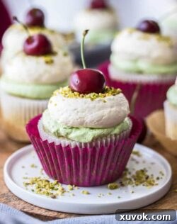 Cherry Pistachio Cupcakes with a bright red cherry on top, featuring two-toned frosting and a visible cherry compote filling.