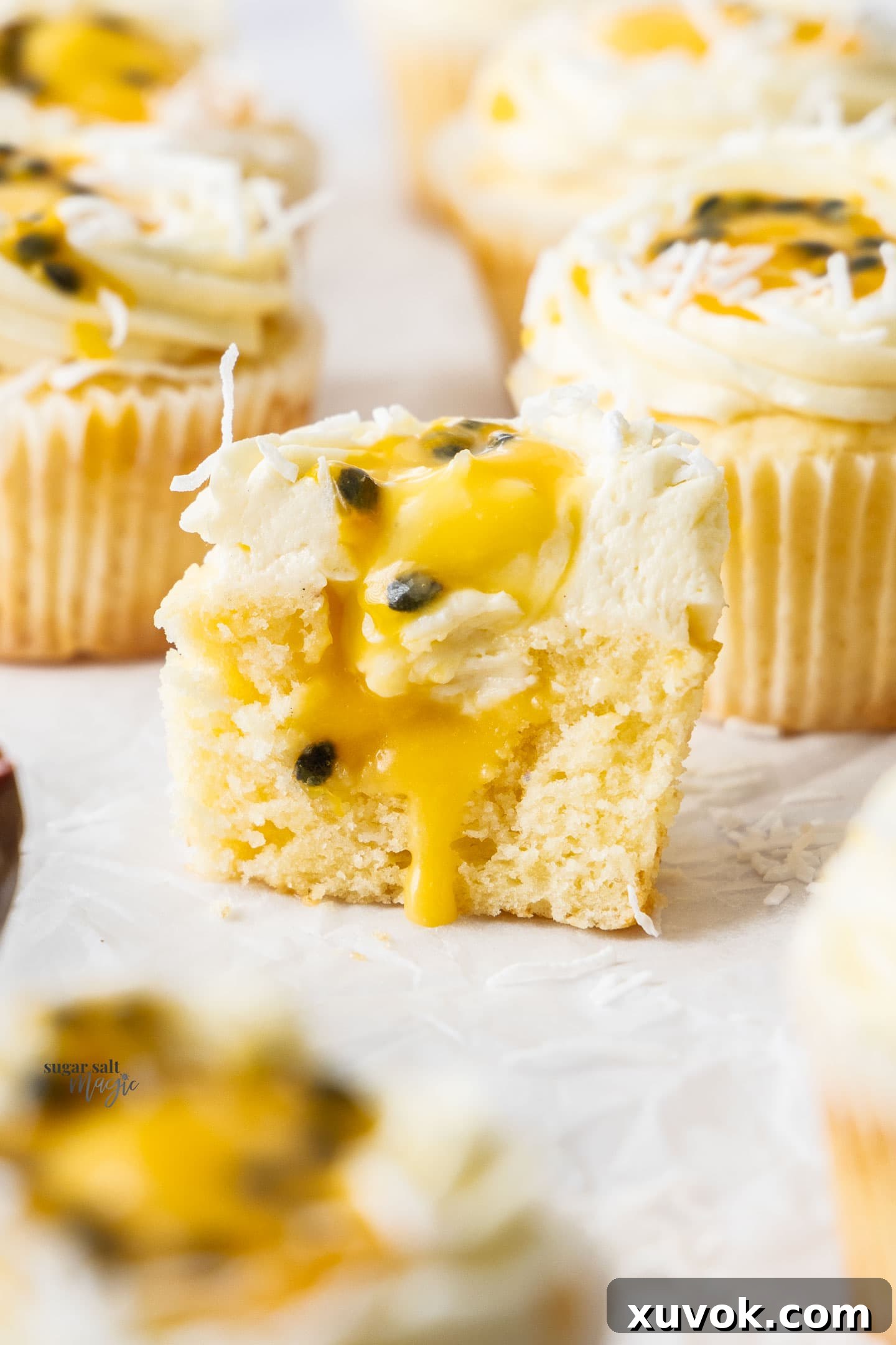 A passionfruit cupcake cut open to show the vibrant passionfruit curd filling inside, topped with coconut buttercream and fresh passionfruit seeds.