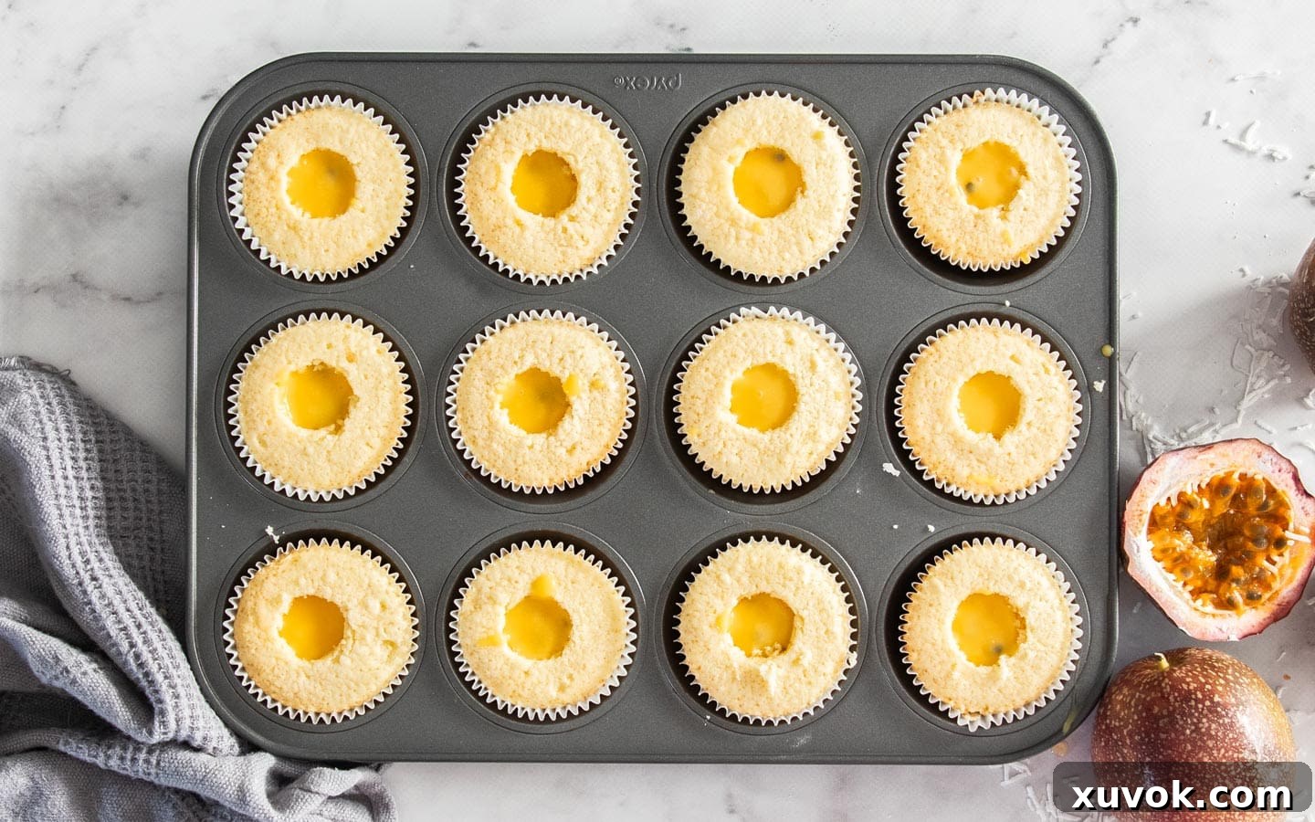 Cooled cupcakes in a muffin pan, with their centers cored out and filled with bright yellow passionfruit curd.