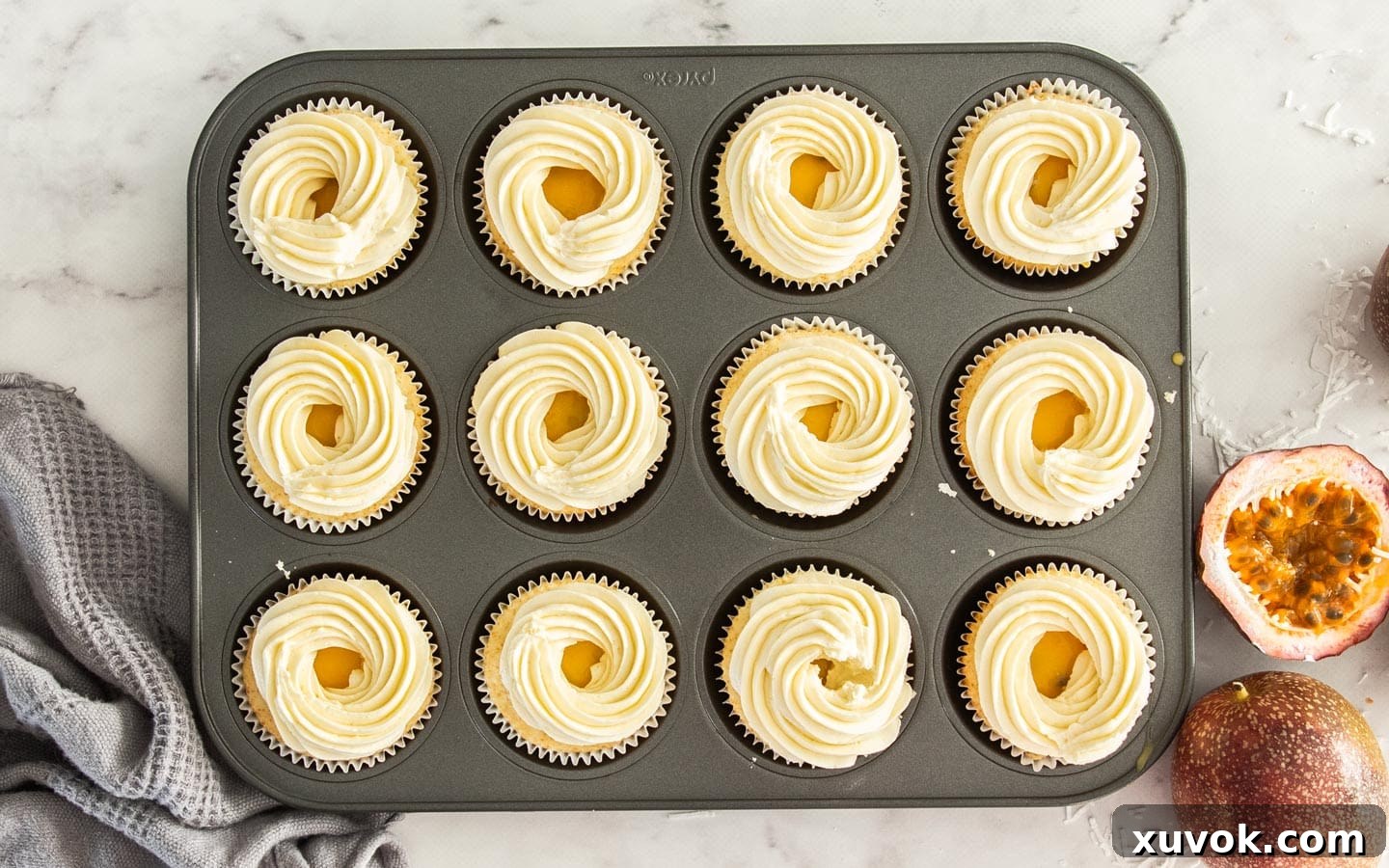 Cupcakes in a muffin pan, each topped with a swirl of coconut buttercream, covering the curd filling but leaving a small indentation in the center.