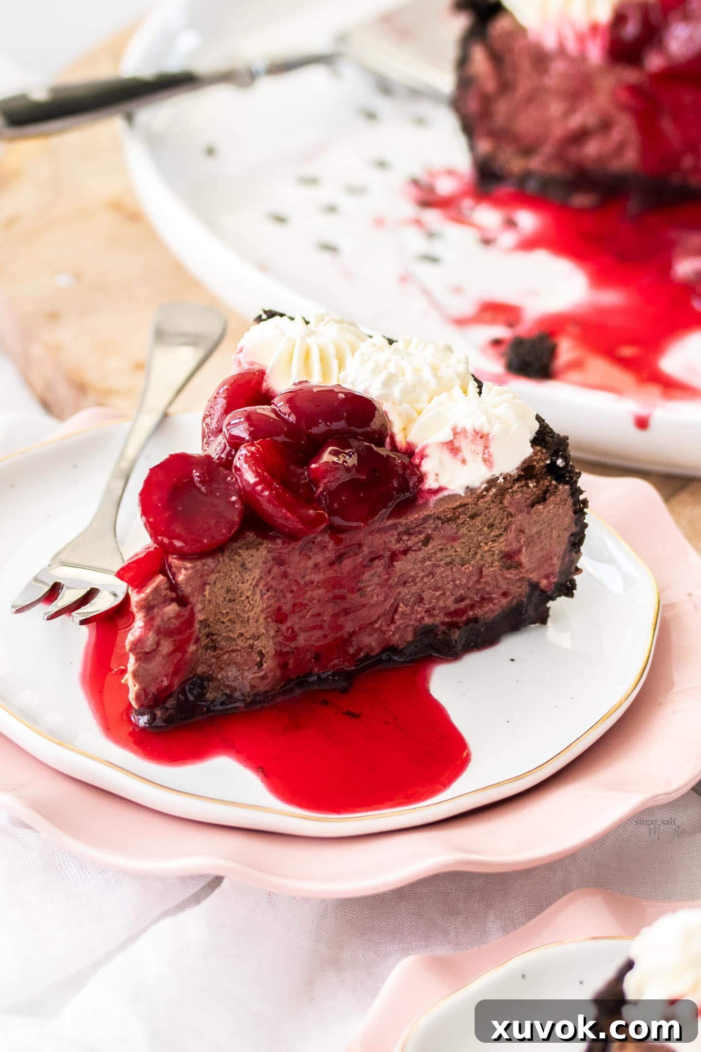 A perfectly portioned slice of Black Forest Cheesecake on a white dessert plate, showing its layers of dark crust, chocolate filling, whipped cream, and cherry sauce.