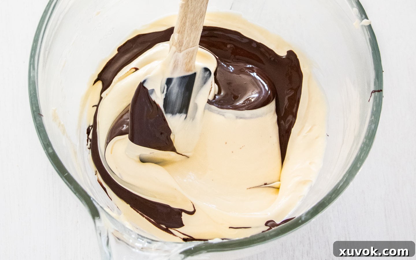 Melted dark chocolate being carefully folded into the light-colored cheesecake batter with a spatula.