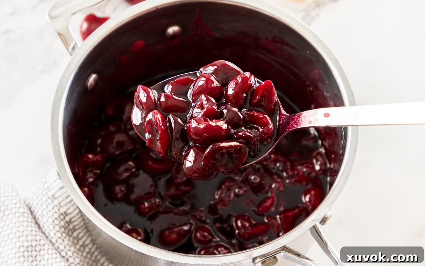 A scoop of rich, dark red cherry sauce being lifted from a saucepan, showing its thick and glossy texture.