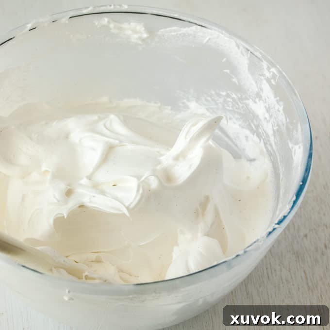 A pristine, glossy meringue (marshmallow frosting base) in a clear glass bowl, showcasing its thick, stable, and perfectly whisked texture, ready for flavoring.