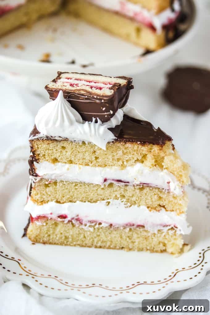 A close-up, inviting image of a perfectly sliced Wagon Wheel Cake on a pristine white plate, showcasing its distinct layers of vanilla sponge, vibrant raspberry jam, and fluffy marshmallow frosting, all crowned with chocolate ganache and mini Wagon Wheel pieces.