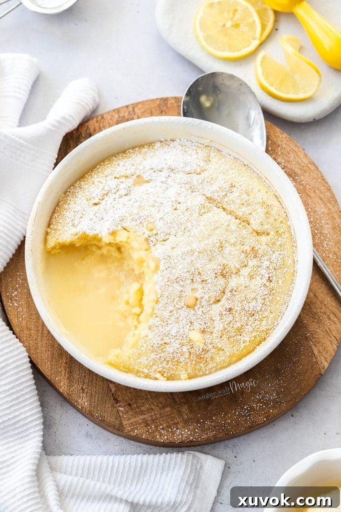 Top down view of a warm self-saucing lemon pudding in a round white bowl, with a scoop taken out, and lemons in the corner.