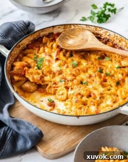 A white pan filled with creamy red pasta bake. A grey tea towel sits next to it. A wooden spoon on top.